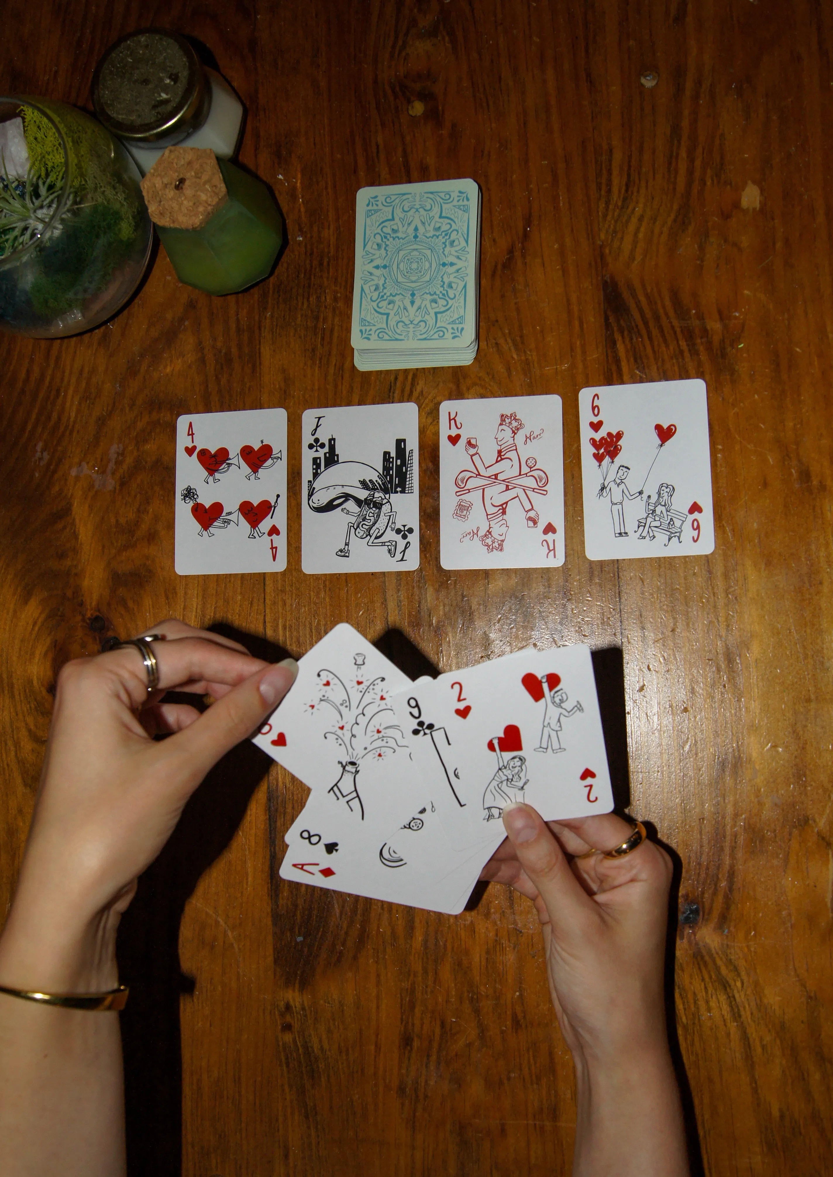 Hands holding a fan of the custom cards over a wooden table, with four illustrated cards laid out above them and the patterned deck stacked at the top.