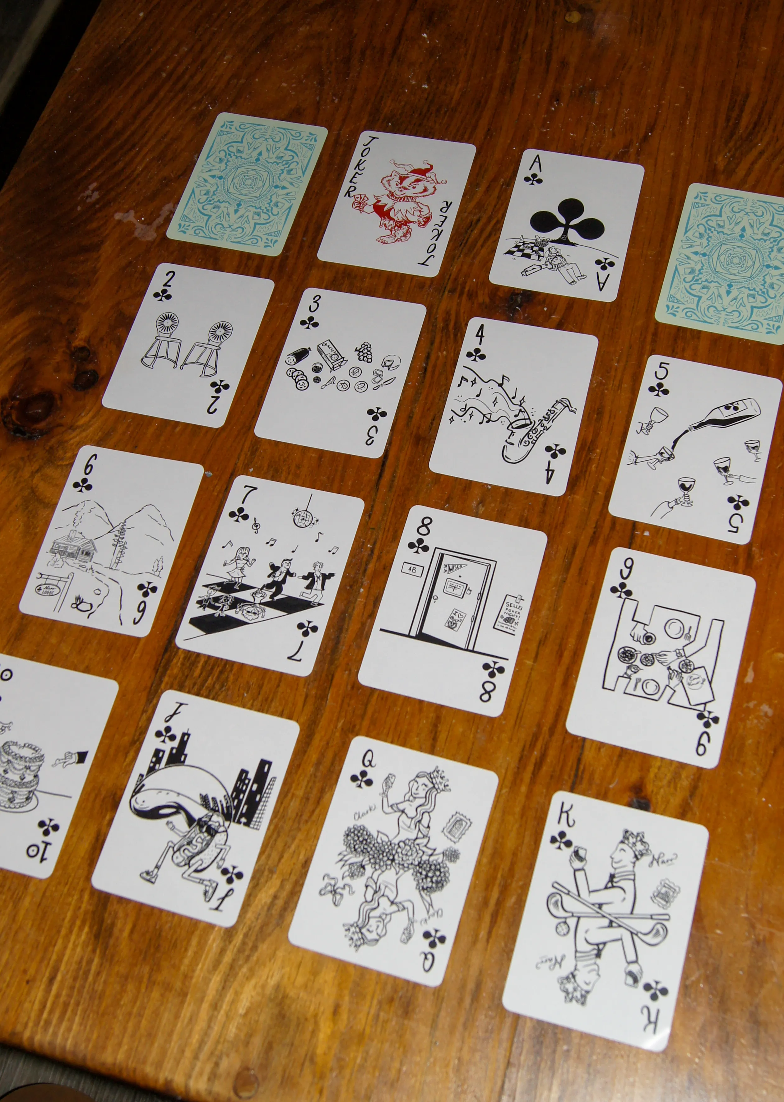 The full suit of clubs laid out in rows, illustrated in black — bar stools, a saxophone, a disco dance floor, a tiered wedding cake, and the Joker drawn as Bucky Badger.