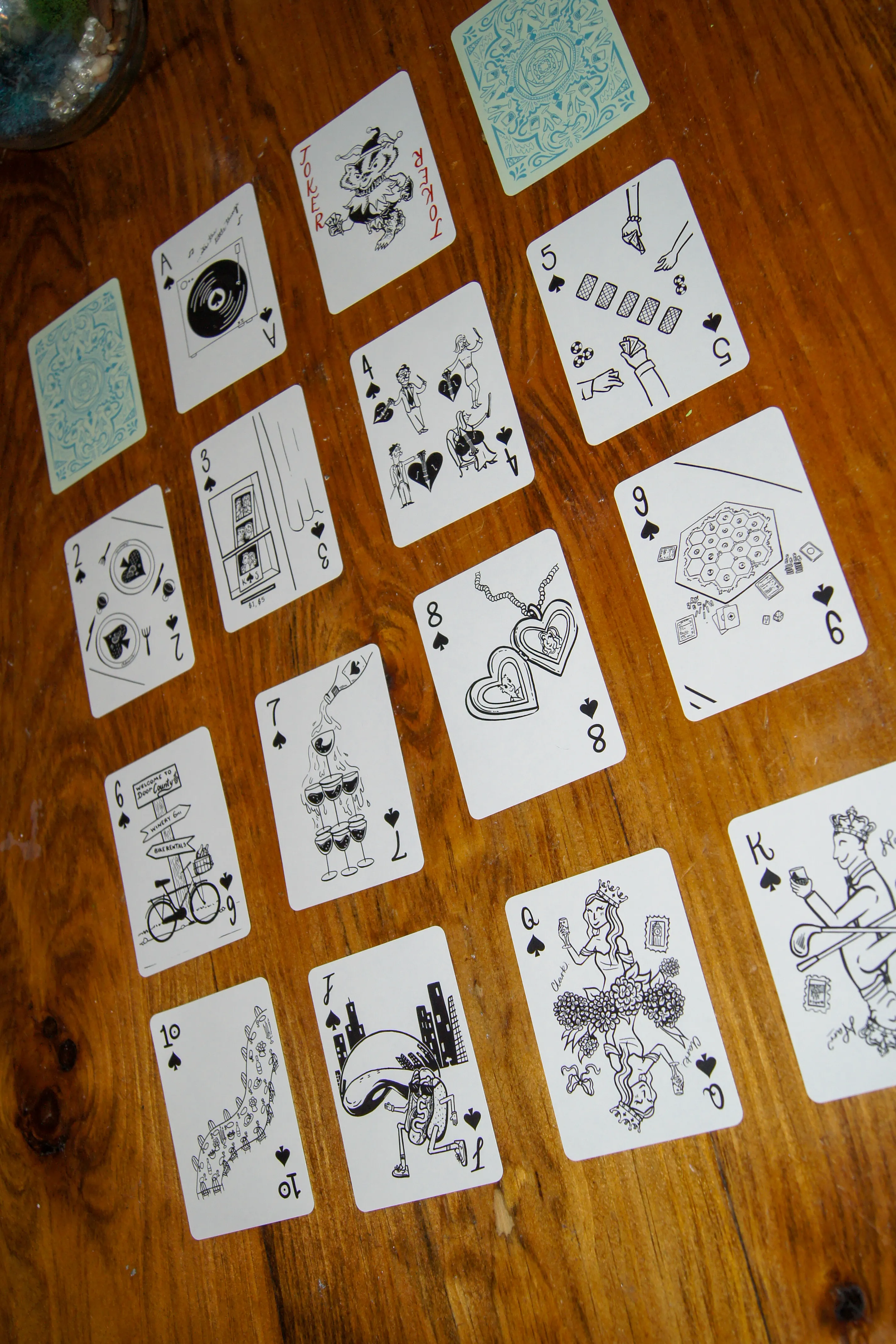 The full suit of spades laid out in rows, illustrated in black — a record player, a heart locket, a board game night, and a bicycle at a Door County signpost.